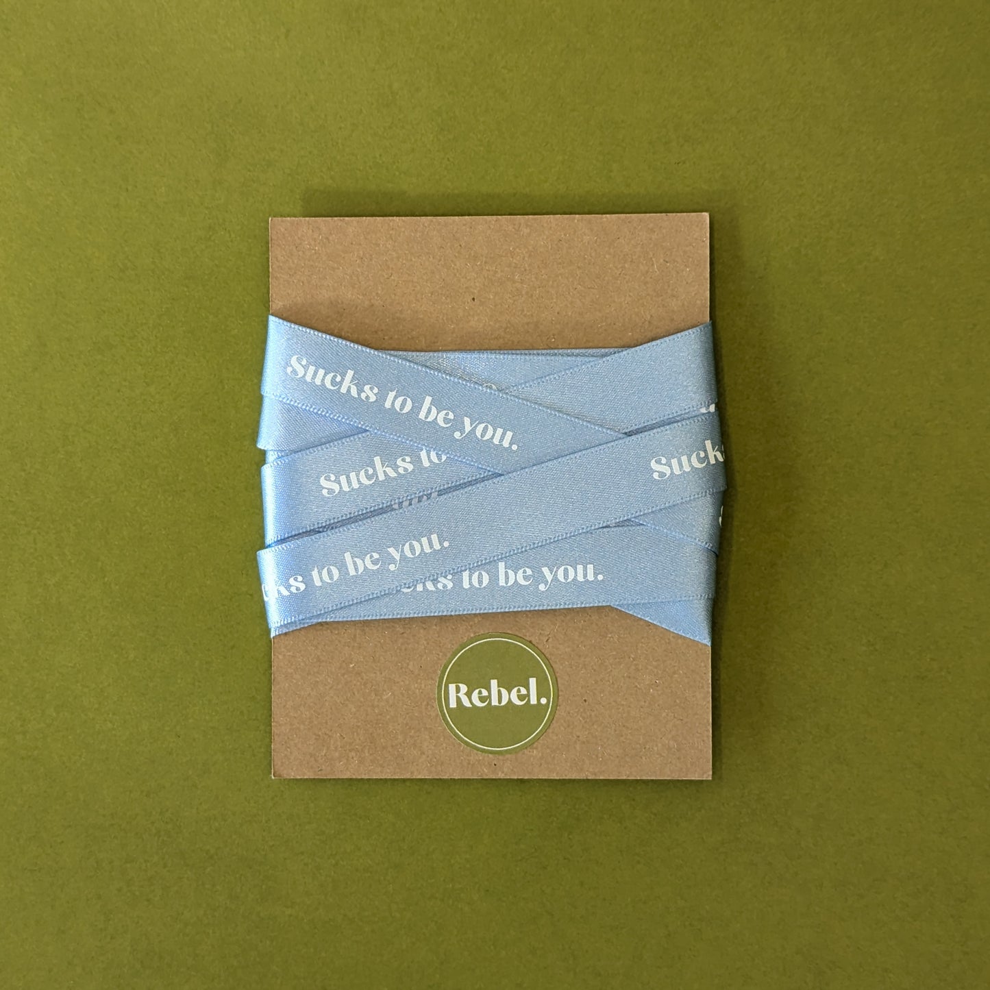 A blue satin ribbon with the phrase 'Sucks to be you' printed on it, presented on a recycled Kraft board packaging.