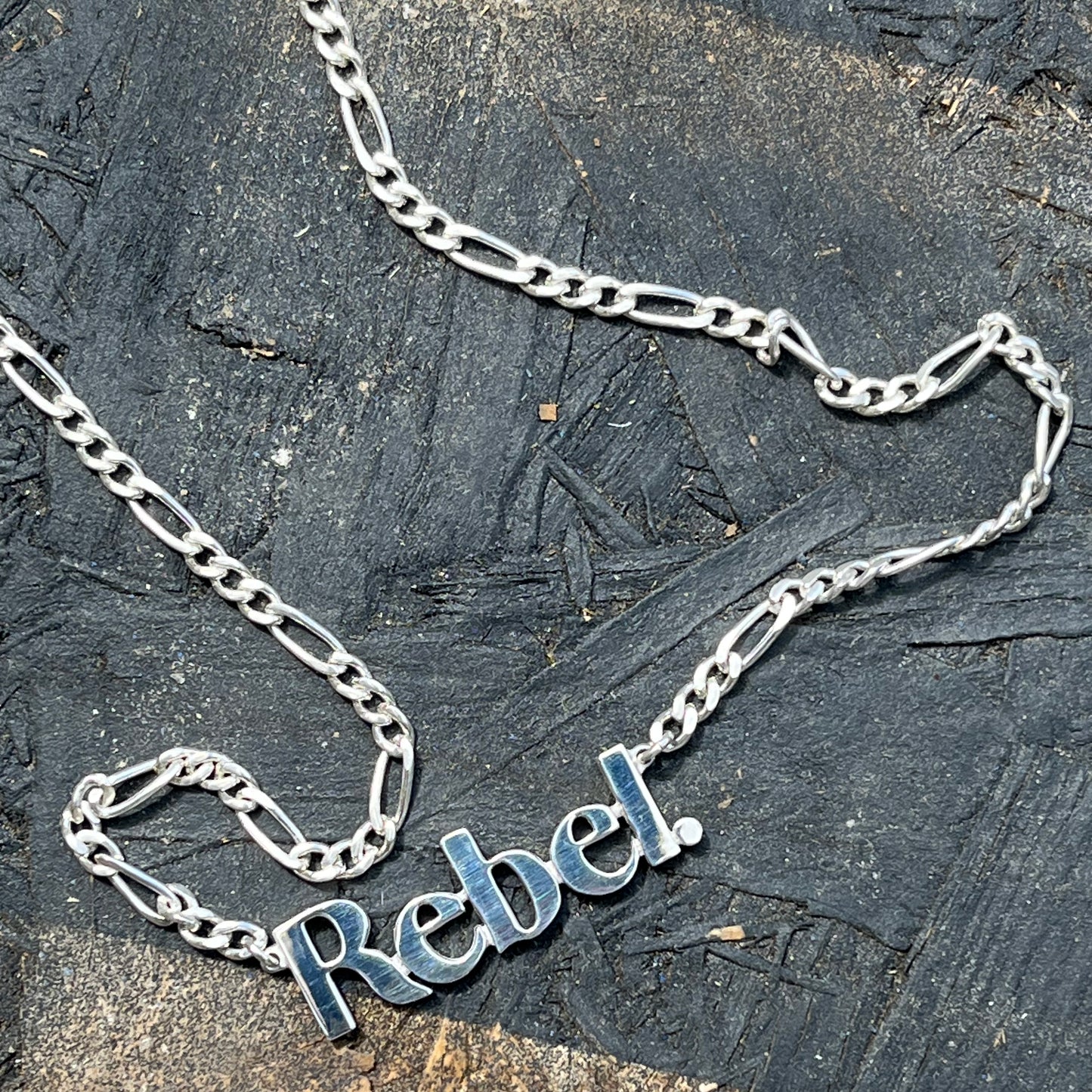 Silver chain necklace with 'Rebel' pendant on a textured dark surface