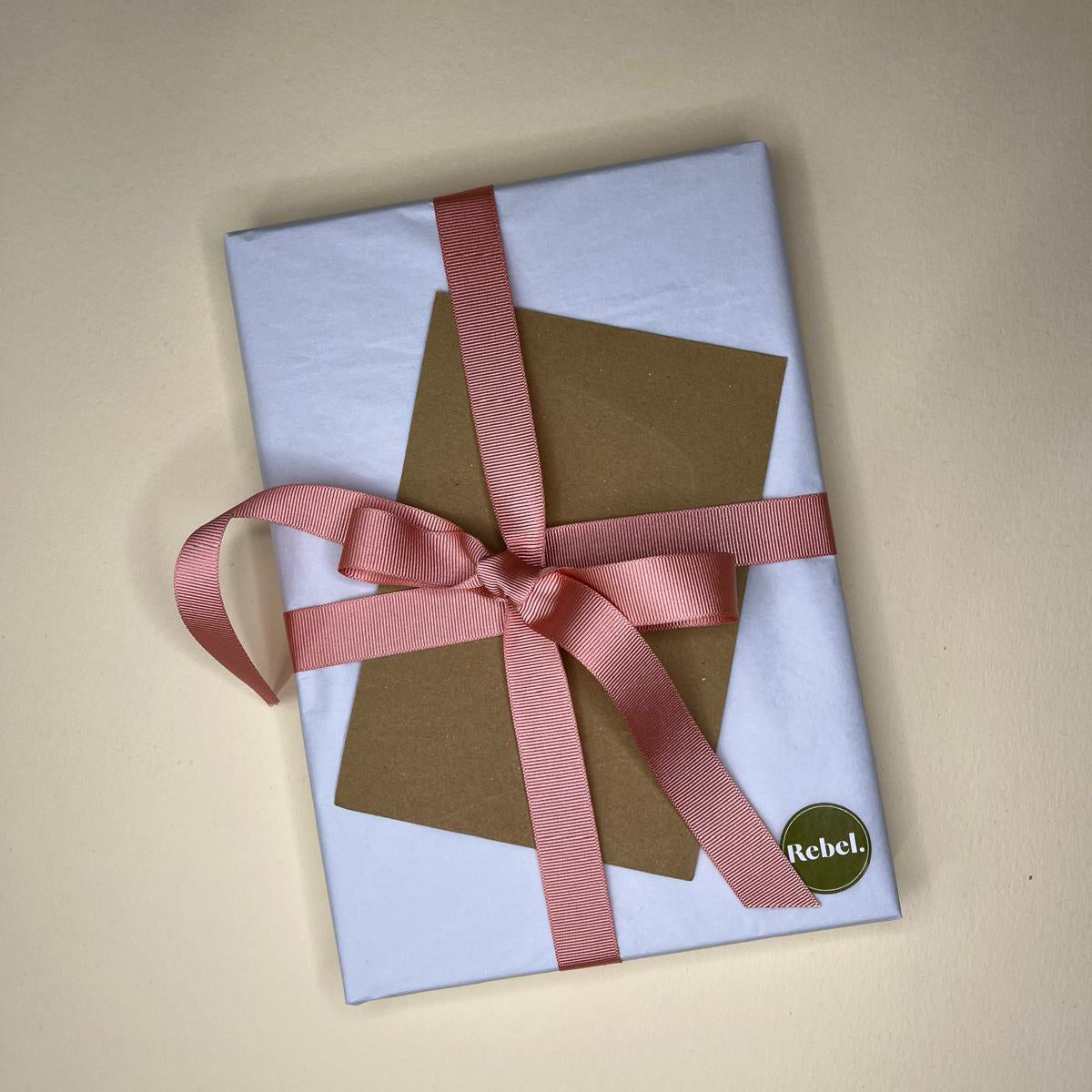 A5 Notebook wrapped in white tissue paper with pink grosgrain ribbon, brown card and green Rebel sticker on beige background