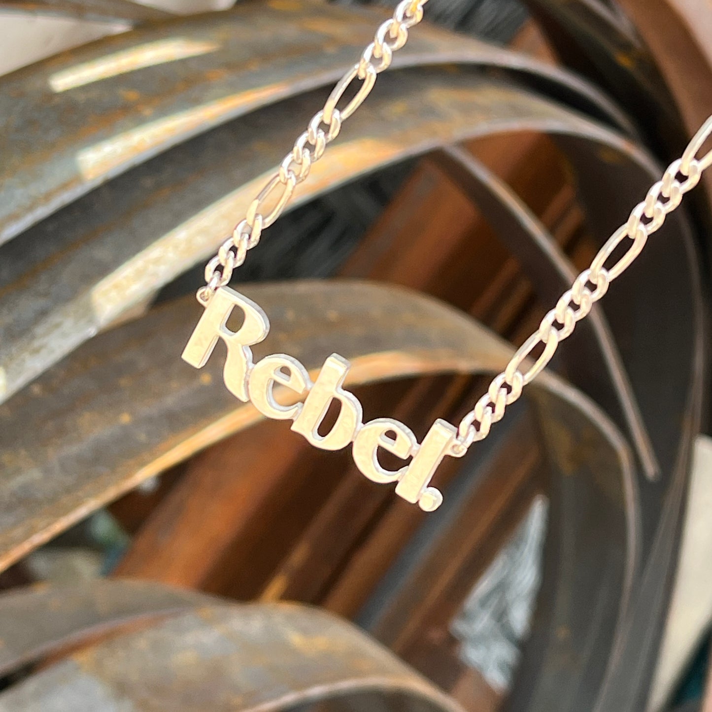 Silver necklace with 'Rebel' pendant on a textured surface