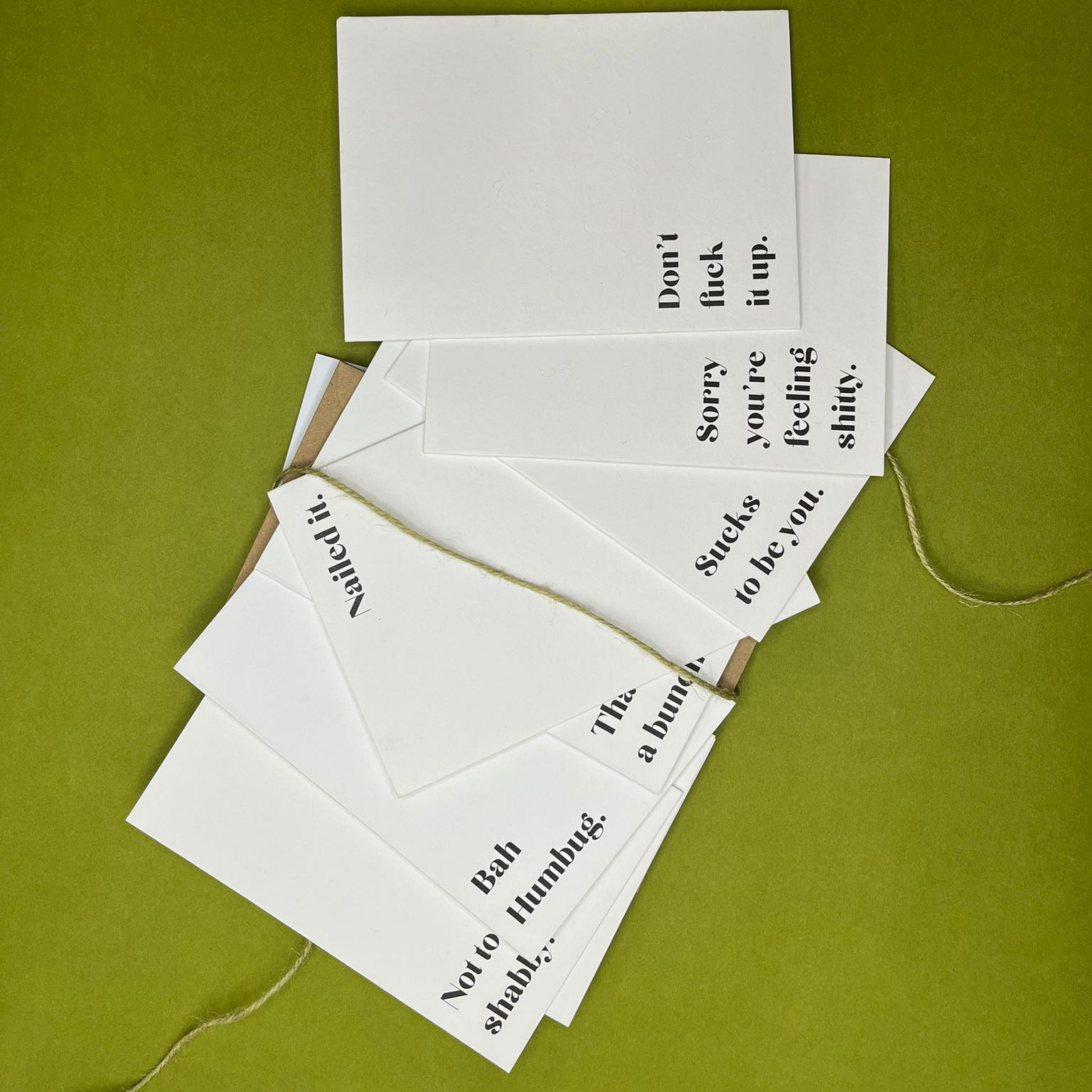 Bundle of white greeting cards with black text spread out with green twine and Kraft envelope on green background